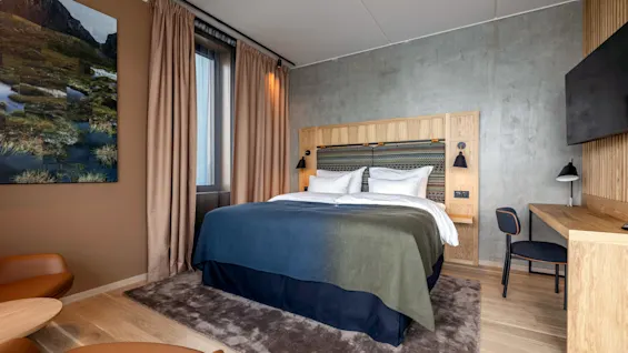 Seng med blågrønt tæppe i moderne hotelværelse; omgivet af træmøbler, brune gardiner, landskabsfotografi på væggen og skrivebord med stol og lampe; beige og betonvægge.