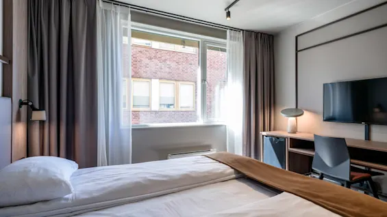 En seng der er redt og står vendt mod et vindue med gardiner i et hotelværelse. Et lille skrivebord og stol står ved siden af under et vægmonteret TV.