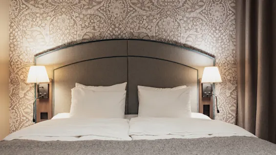 En bekväm hotellsäng med grå sänggavel, vita sängkläder och varma vägglampor mot mönstrad tapet.