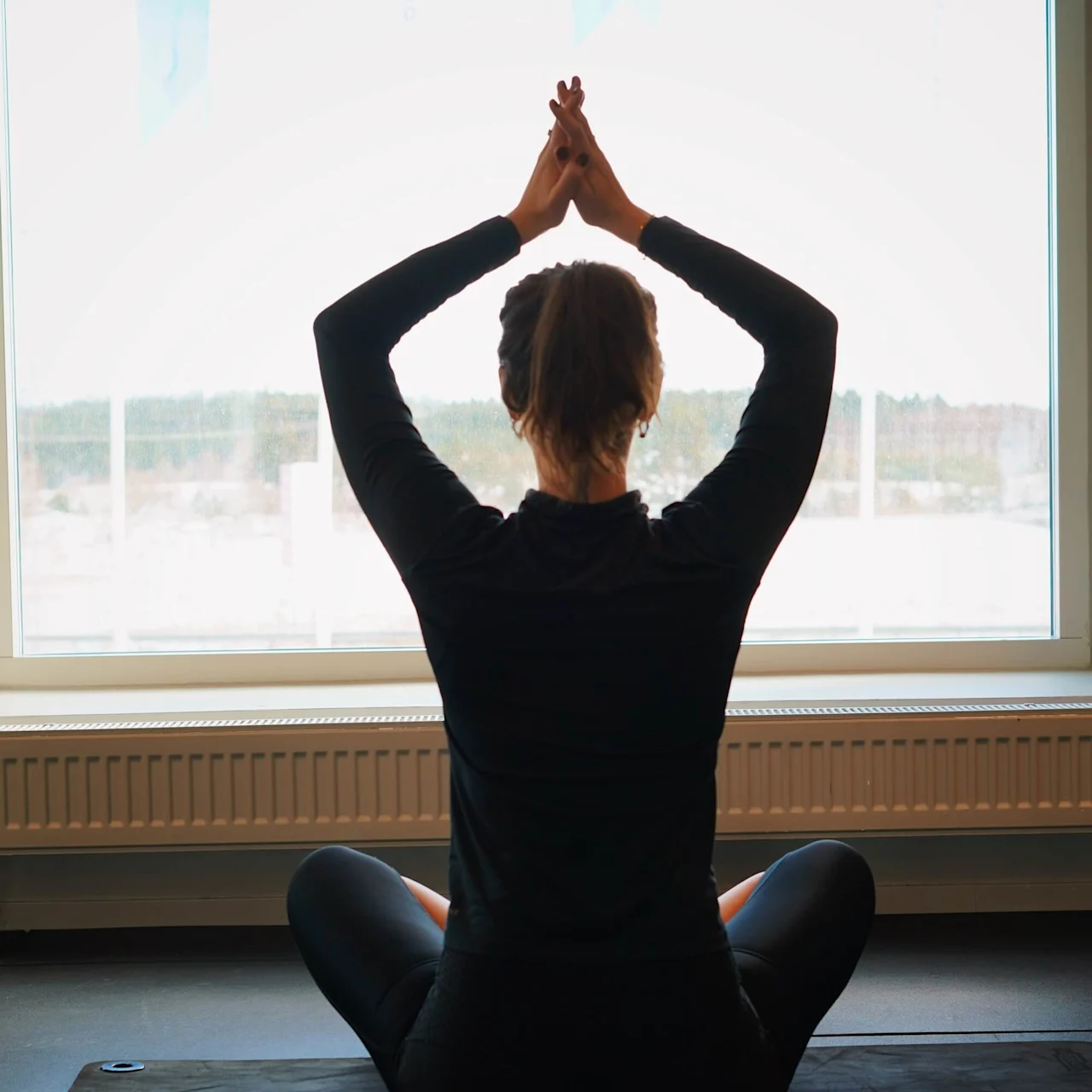 Person sitting cross-legged on a yoga mat, hands raised above head, in front of a large window with a snowy landscape outside.