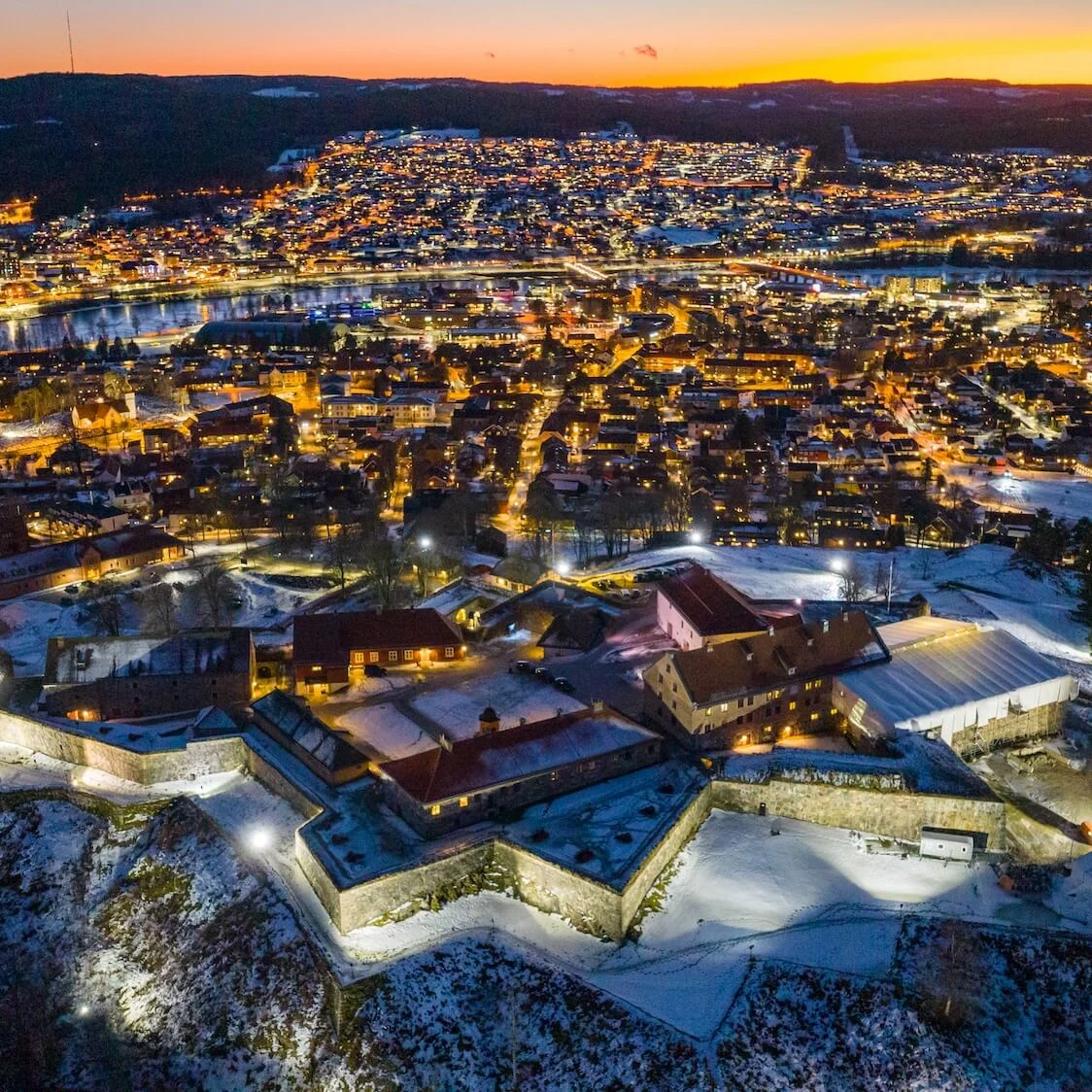 En befæstning på en bakke med stenmure er oplyst i skumringen. Det har udsigt over en oplyst byscene med bygninger, veje og en flod, der reflekterer solnedgangen.