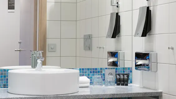 En badrum har ett vitt handfat på en kaklad bänk, med en rund spegel och handdukshyllor ovanför. Två svarta behållare för bekvämligheter bredvid kranen i en kompakt, modern miljö.