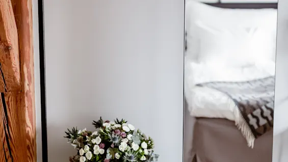Et smart soveværelsesarrangement med en mørk trækommode, et levende blomsterarrangement, et tøjstativ og et spejl, der reflekterer en behagelig seng.