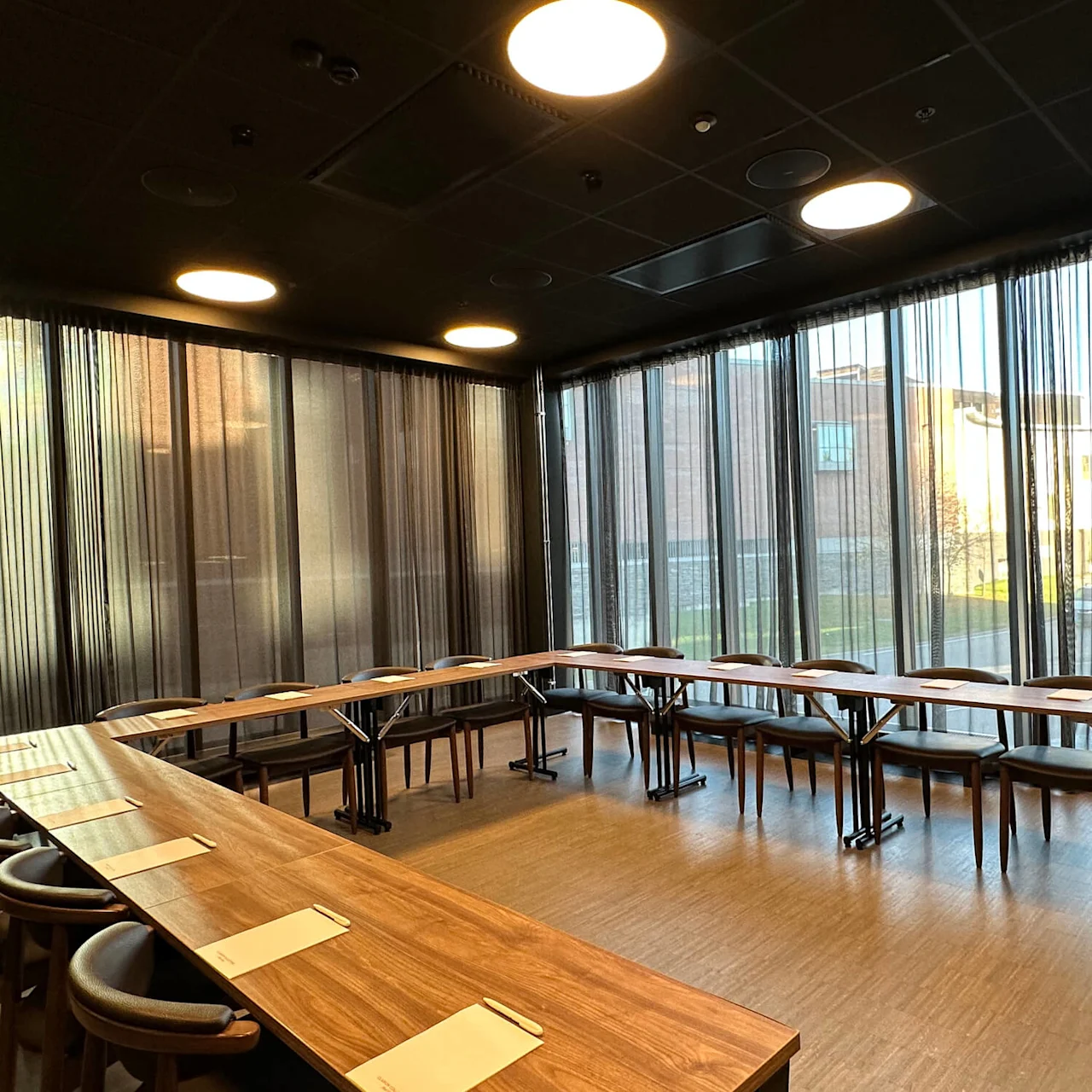 Conference tables with chairs in a modern room, sunlight filtering through vertical blinds, empty and quiet.