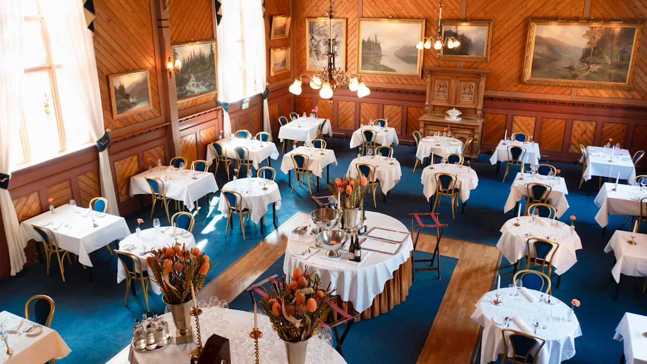 Elegant dining tables, set with white tablecloths, fill a richly decorated room with wooden paneling and artwork. Warm lighting from chandeliers creates an inviting, classic ambiance.