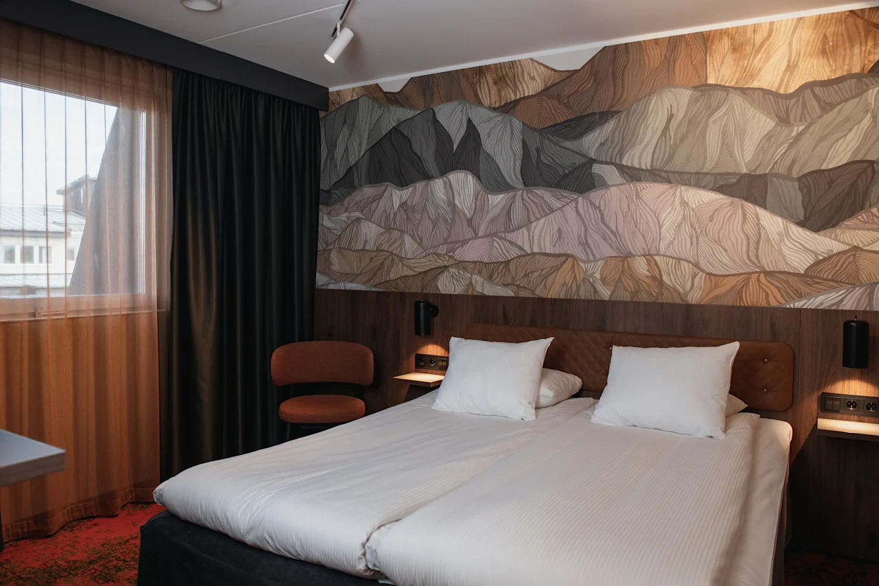 Et koselig hotellrom med en stor seng, et vindu med gardiner og en dekorativ vegg med et fjellandskapsdesign.