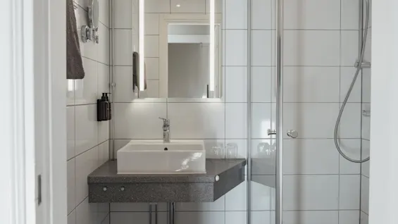 Puhdas ja moderni kylpyhuone Home Hotel Victoriassa, jossa on valkoinen pesuallas, valaistu peili ja lasinen suihkukaappi.