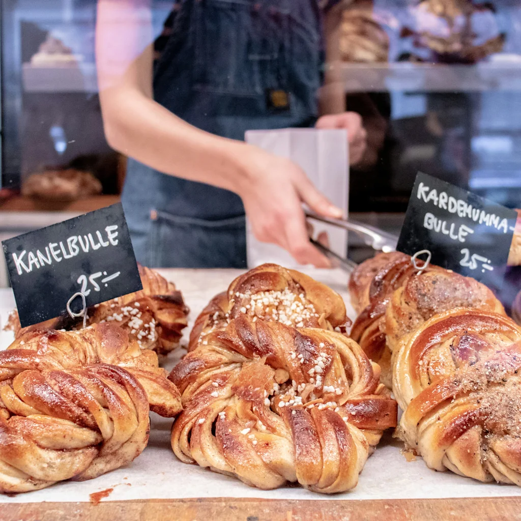 En person serverar kanelbulle och kardemummabulle i ett bageri, perfekt för en svensk fika. Kanelbulle 25, Kardemummabulle 25.