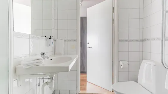 A clean, white-tiled bathroom with a sink, mirror, toilet, and an open door revealing a room with a wooden floor.