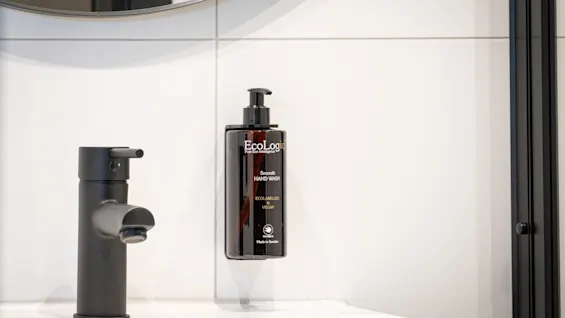 A modern bathroom with a black faucet and a hand wash dispenser on a white tiled wall. Text: Smooth HAND WASH ECOLABELLED VEGAN Made in Sweden.