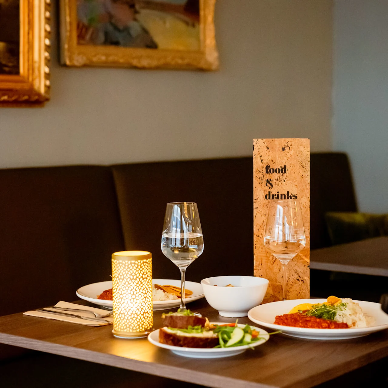 Hyvin katettu ruokapöytä ruoan ja juomien kera, lämmin valo kynttilälyhdystä ja 'Food & Drinks' menu-teline.