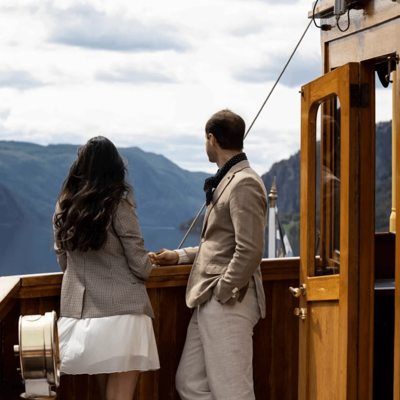 Two people stand on a wooden boat deck overlooking a calm fjord, surrounded by forested mountains under a cloudy sky.