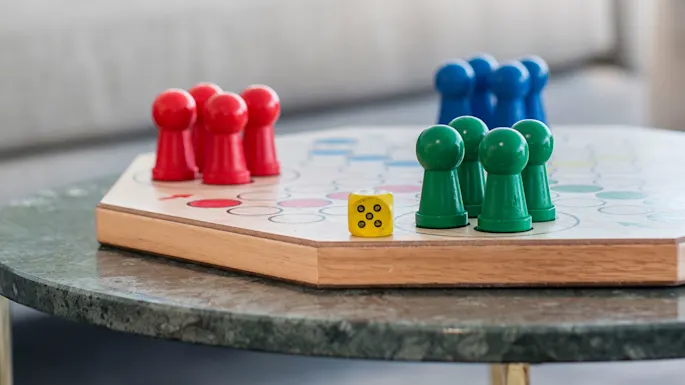 Brettspill med fargerike brikker og en terning står på et rundt bord; uskarp sofa med puter i bakgrunnen.