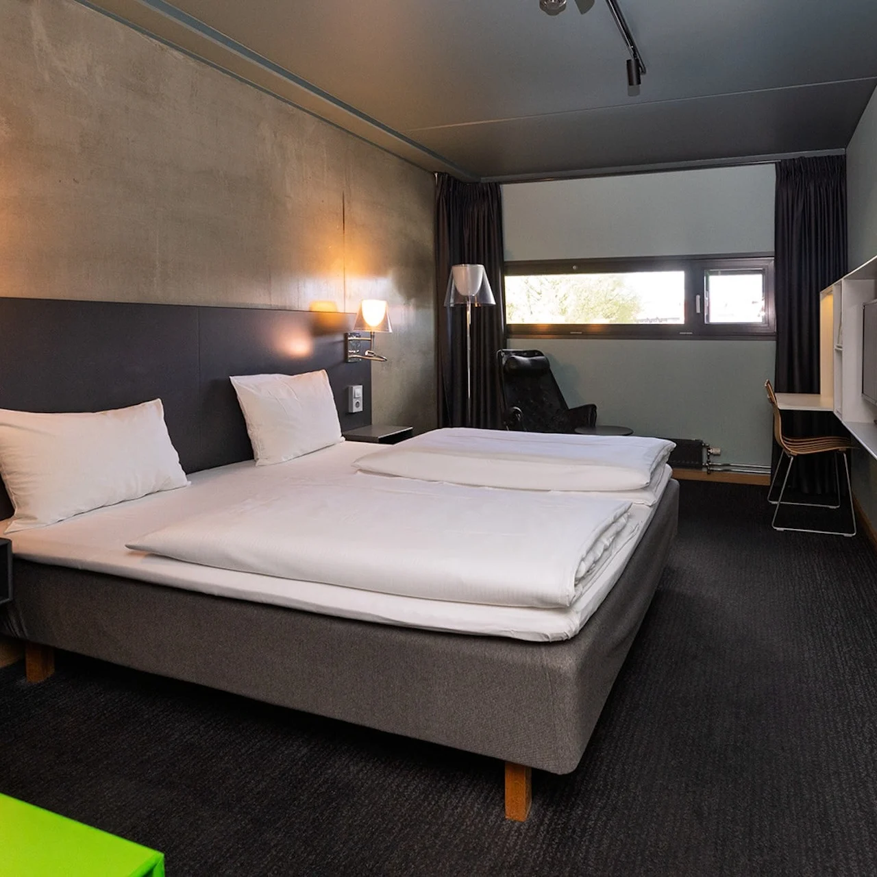 A neatly made double bed with white linens stands in a modern hotel room, featuring dark walls and a small desk near a window, illuminated by wall-mounted lamps.