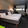 A neatly made double bed with white linens stands in a modern hotel room, featuring dark walls and a small desk near a window, illuminated by wall-mounted lamps.