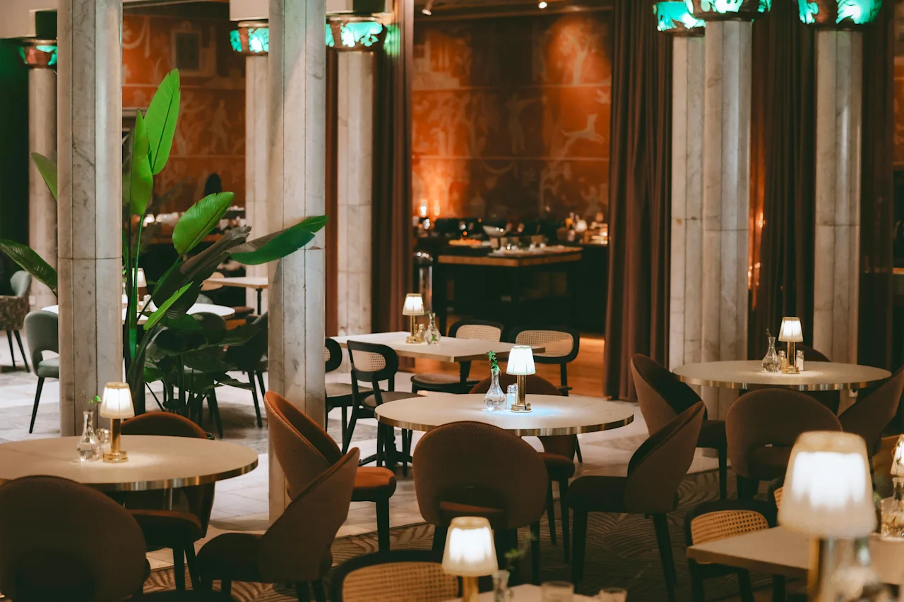 Tables are set with lamps and glasses in a warmly lit restaurant, surrounded by pillars and decorative plants, creating an inviting dining atmosphere. No text is present.