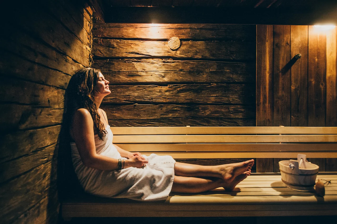 En kvinde, indhyllet i et håndklæde, slapper af på en træbænk i en svagt oplyst sauna, omgivet af rustikke trævægge, med en træspand i nærheden.