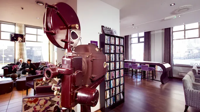 A vintage projector stands in a modern lounge area, where people sit and chat, surrounded by DVDs and urban views through large windows.