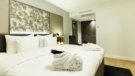 A neatly made bed with folded robes lies in a hotel room featuring floral artwork, bedside lamps, and a full-length mirror near a closed door.