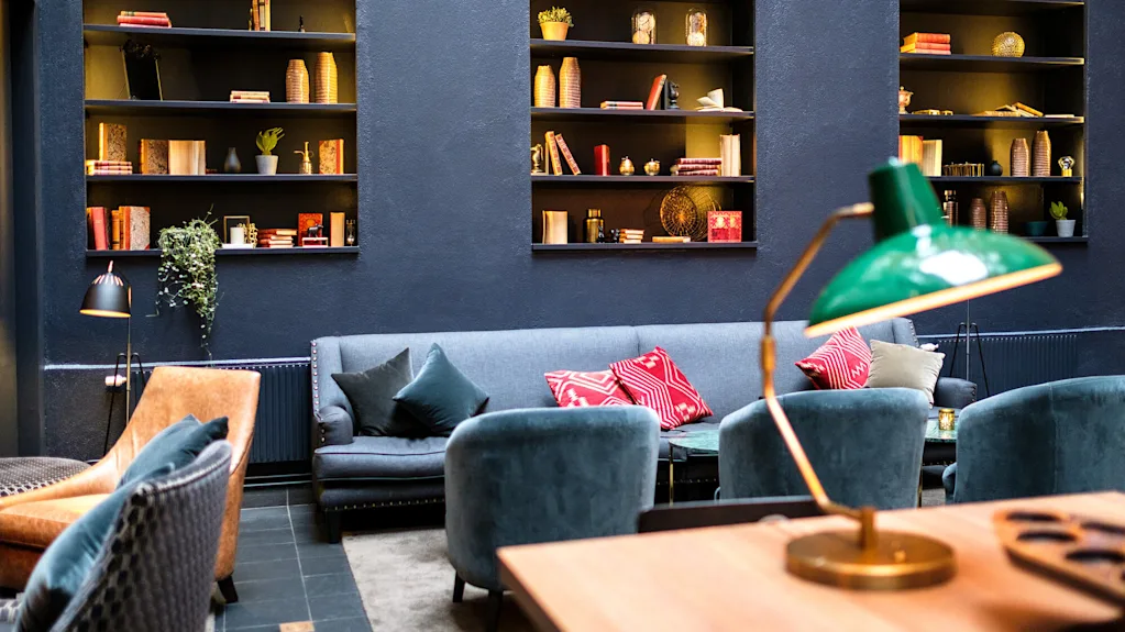 A cozy lounge area with plush sofas, pillows, a bookshelf, and a prominent green desk lamp illuminating a wooden table.