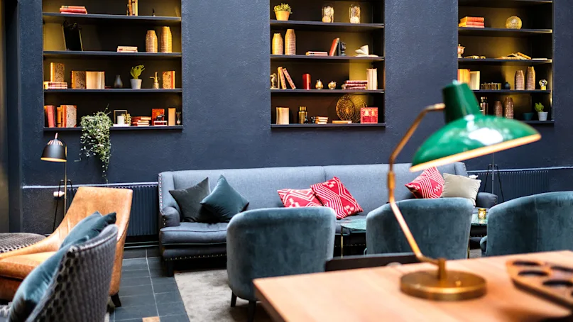 A cozy lounge area with plush sofas, pillows, a bookshelf, and a prominent green desk lamp illuminating a wooden table.