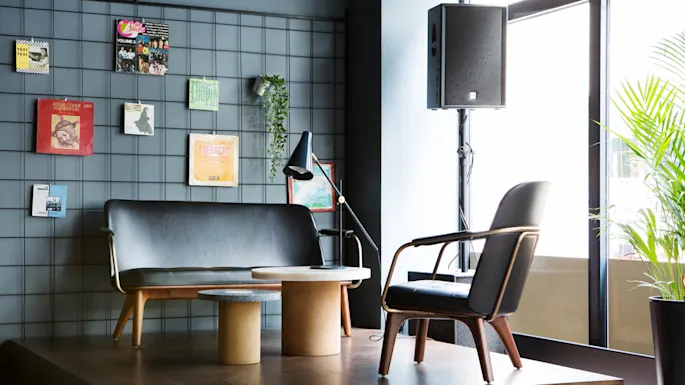 A modern seating area features a black sofa and chair on a wooden platform, with a grid wall displaying albums and a plant nearby, under a large window.