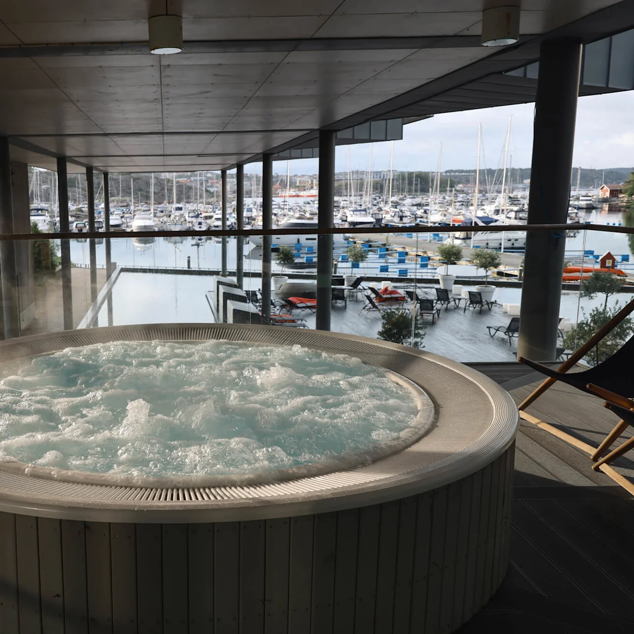 Boblebad bobler aktivt på en overbygd terrasse med utsikt over en marina fylt med båter og omgitt av grøntområder. Et skilt lyder, "Massasjebasseng. Ikke egnet for barn under 5 år."