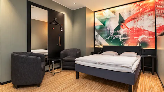 Modern hotel room featuring a double bed, two armchairs, and a colorful artwork on the wall.