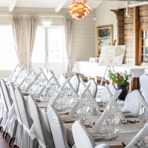 Elegantly set tables with white cloths and napkins await guests in a bright, well-lit room featuring wooden walls, large windows, and decorative lighting fixtures.