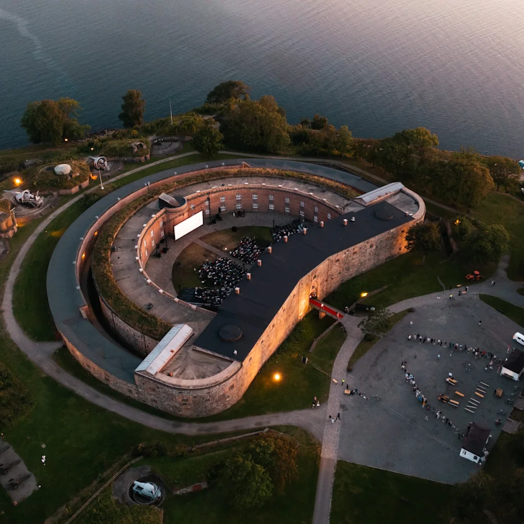 Aerial view of a historic C-shaped building by the water, hosting an outdoor event with a large screen and an audience.