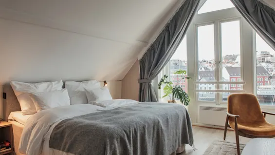 Et hyggeligt soveværelse med en stor seng, en komfortabel lænestol og en plante, med et vindue der vender ud mod en by og havn.