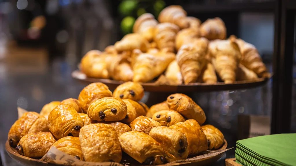 Pastries displayed on wooden platters at a bakery; chocolate croissants are stacked in the foreground, with plain croissants in the background. Green napkins are stacked neatly nearby.
