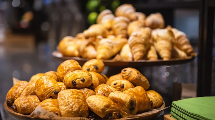 Pastries displayed on wooden platters at a bakery; chocolate croissants are stacked in the foreground, with plain croissants in the background. Green napkins are stacked neatly nearby.