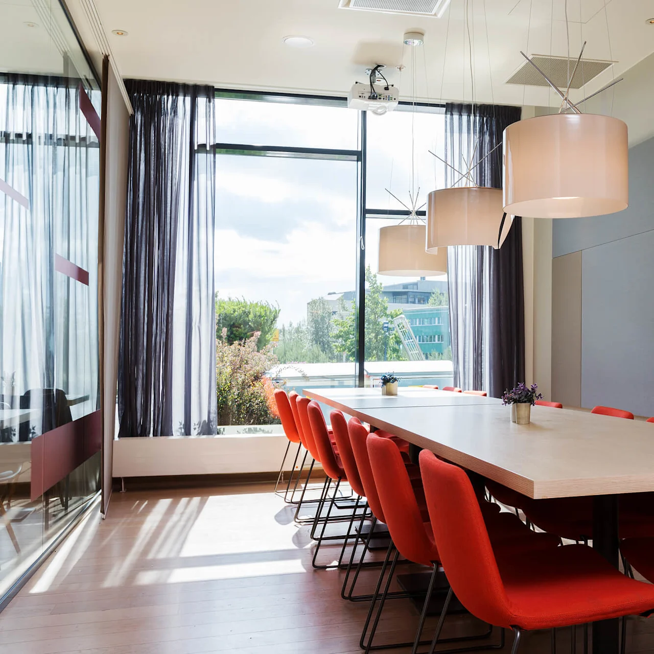 Large wooden conference table surrounded by red chairs under hanging lamps, situated in a well-lit room with large windows and gray acoustic panels.