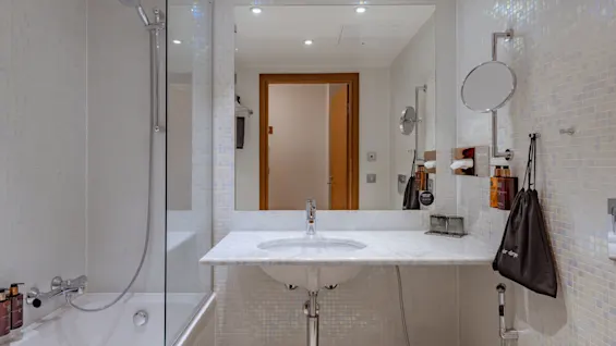 Badeværelse med brusebad med glasvæg, hvide flisevægge, et stort spejl, håndvask og toiletartikler. "Air dryer" pose hænger nær et vægmonteret spejl. Dør reflekteres.
