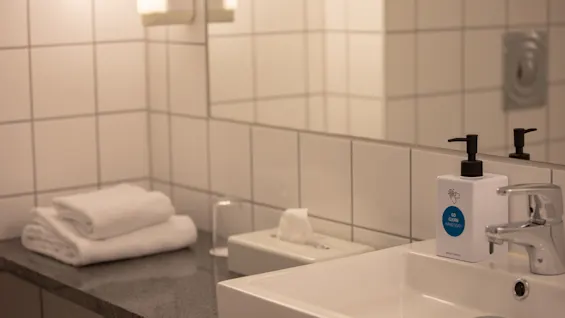 En hvit såpepumpe merket "GO CLEAN HAND SOAP" står på en flislagt baderomsbenk ved siden av en vask, med håndklær og vev i nærheten.