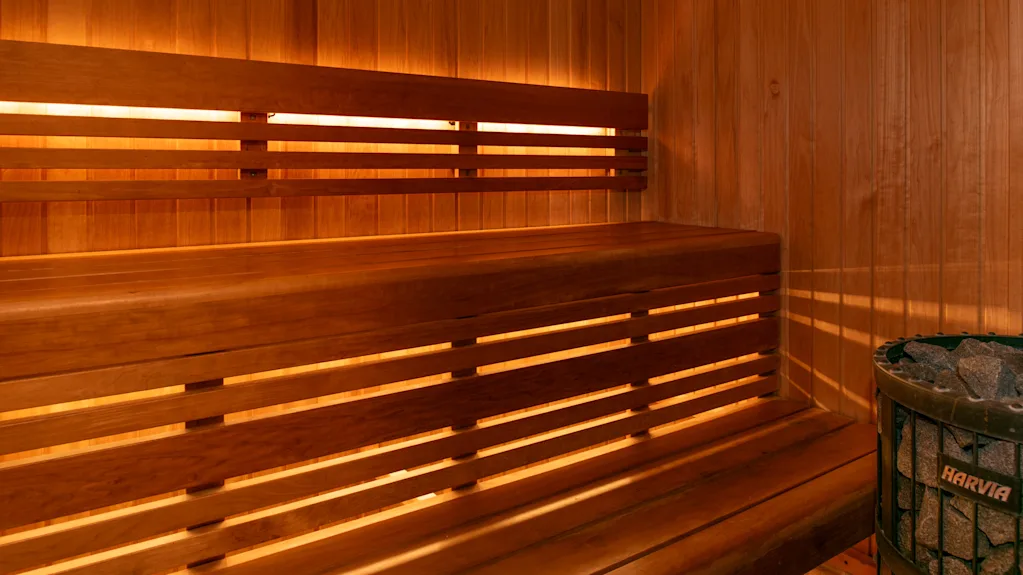 Puistekropolttuinen sauna, jossa on kaksi tasoa penkkejä seinien varrella. Himmennetty valaistus korostaa lämmintä, kodikasta tunnelmaa. Metallinen kiuas kivineen nurkassa.