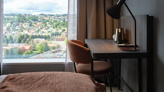 Skrivebord med en lampe og en elkedel står ved siden af et vindue med udsigt over en by. En stol står ved skrivebordet, og en seng er tæt på.