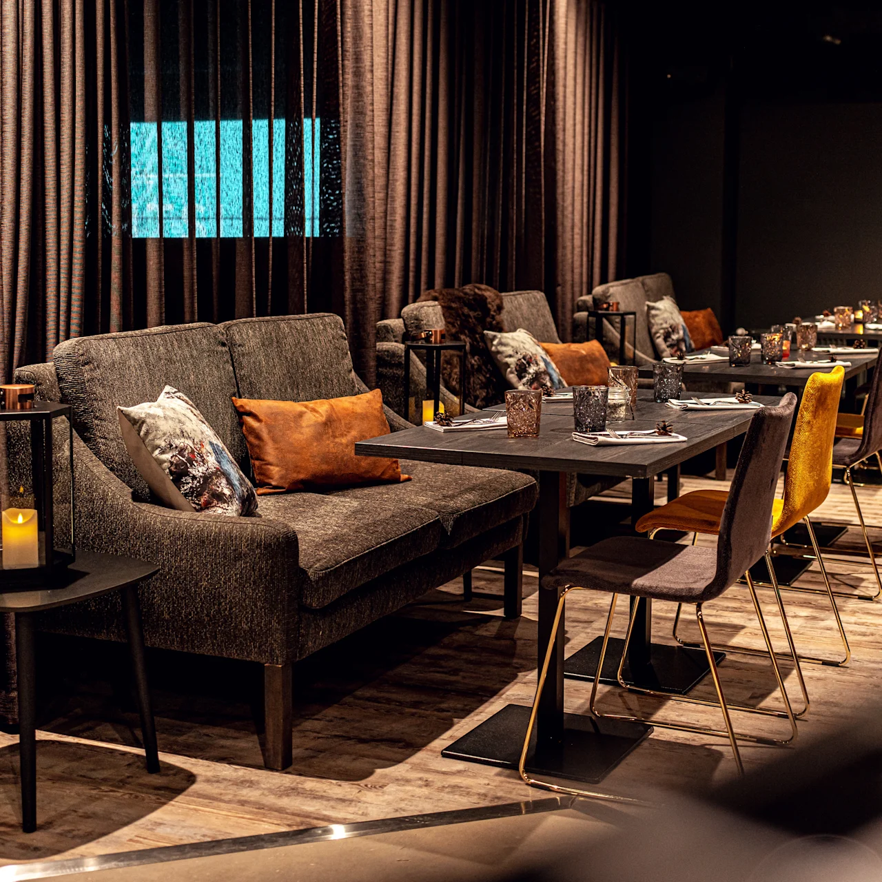 Sofas and chairs surround dining tables with plates and glasses in a dimly lit, cozy room featuring warm brown tones and a candle on a side table.