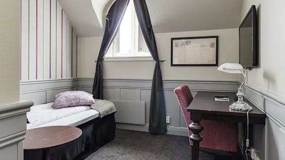 A neatly arranged room with a single bed, window with curtains, desk, chair, and lamp. The decor includes striped wallpaper and framed artwork.