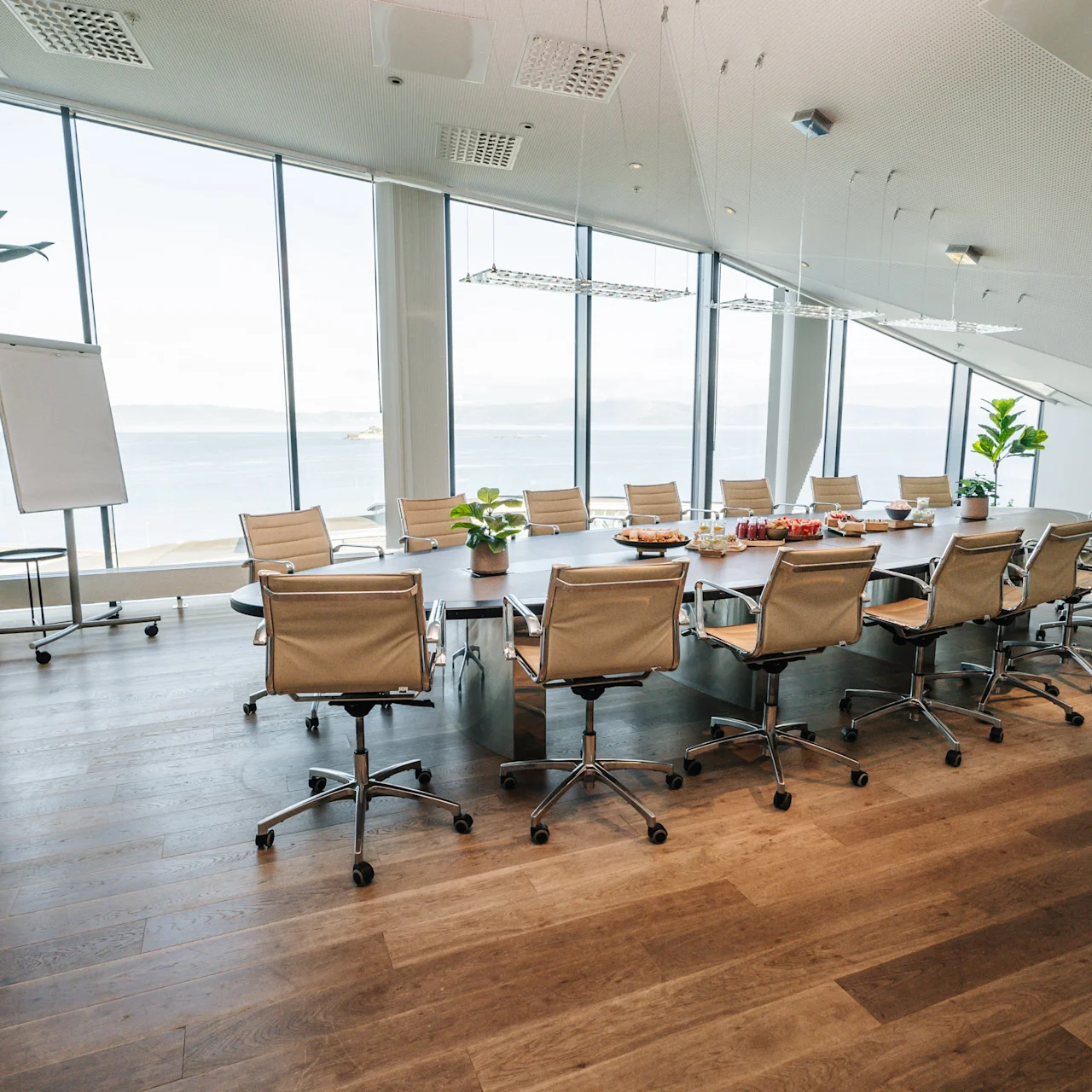 A long conference table with beige chairs surrounds it. Large windows reveal a scenic ocean view. Plants and a flip chart stand accent the modern, airy office space.
