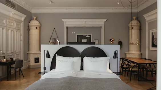 En prydligt bäddad säng med vita sängkläder och grå filt är placerad i mitten av ett elegant rum med två utsmyckade keramiska kaminer och trämöbler, vilket skapar en klassisk, sofistikerad atmosfär.