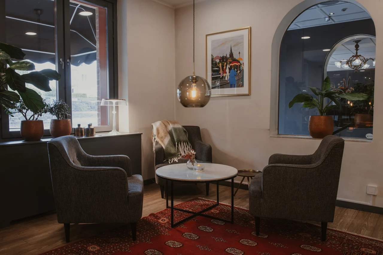 A warm and inviting hotel lounge with comfortable armchairs, a coffee table, and green plants by the windows. Add Home Hotel Kung Oscar.