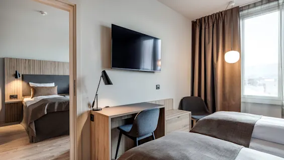 Et moderne hotelværelse har to senge, et vægmonteret TV over et skrivebord med lampe og stol, samt et vindue med brune gardiner, der slipper dagslys ind.