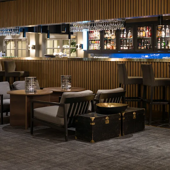 Bardisken viser flere flasker sprit; barstoler står langs disken mens bord med moderne stoler fyller det teppelagte loungeområdet, og skaper en elegant og koselig atmosfære.