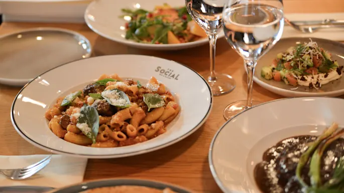 A pasta dish garnished with herbs sits on a plate labeled "SOCIAL." Surrounding dishes and two wine glasses suggest a restaurant table setting with various foods.