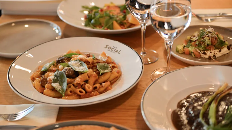 A pasta dish garnished with herbs sits on a plate labeled "SOCIAL." Surrounding dishes and two wine glasses suggest a restaurant table setting with various foods.