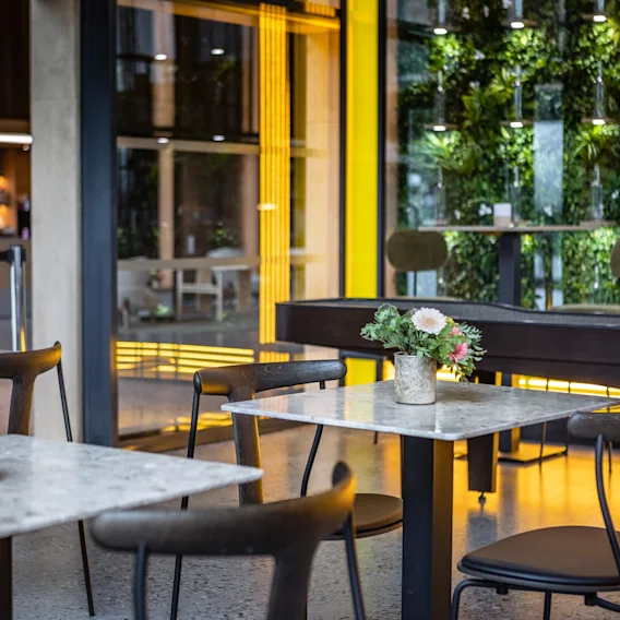 Borde med stoler og blomsterpotter står i en lyst opplyst kafé. En vertikal hage dekorerer veggen, opplyst av gult lys.
