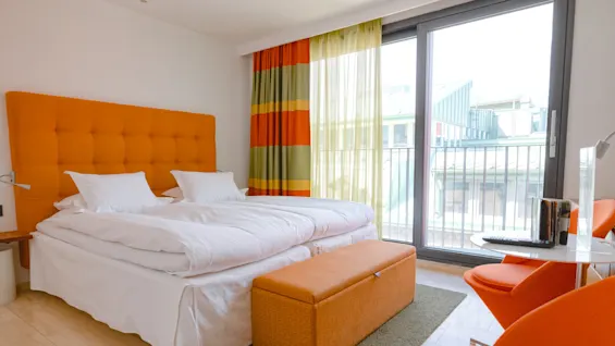 A bed with white linens sits against an orange headboard in a brightly-lit room, featuring patterned curtains, orange furniture, and a large window overlooking a building.