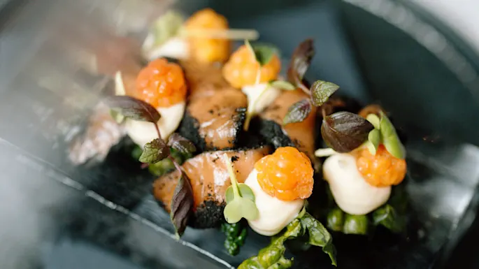 En rett viser delikat anordnet røkt laks toppet med kremklatter, gule bær og mikrogrønt, satt på en glassblokk, servert på en sort tallerken.